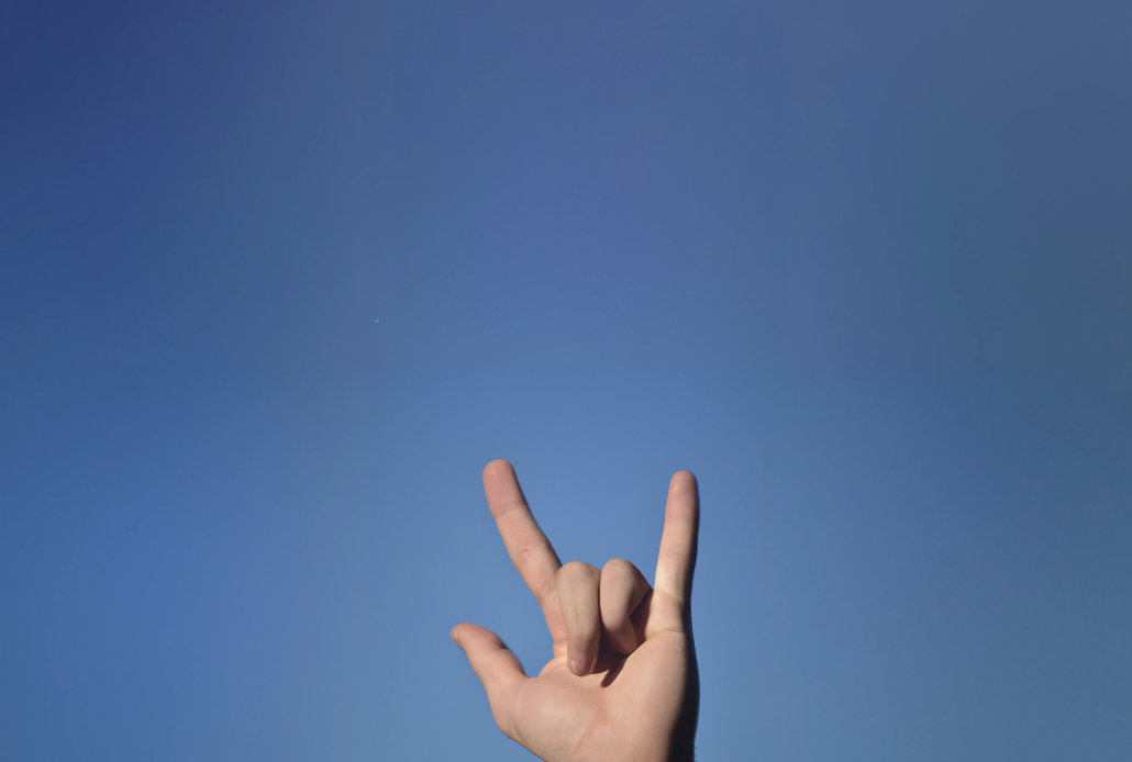 A photograph with the sky in background with no clouds and a hand doing the horn sign, the hand is 3_4 view, the hand is a bit far from the objective, extend the background’s sky on both sides of the picture